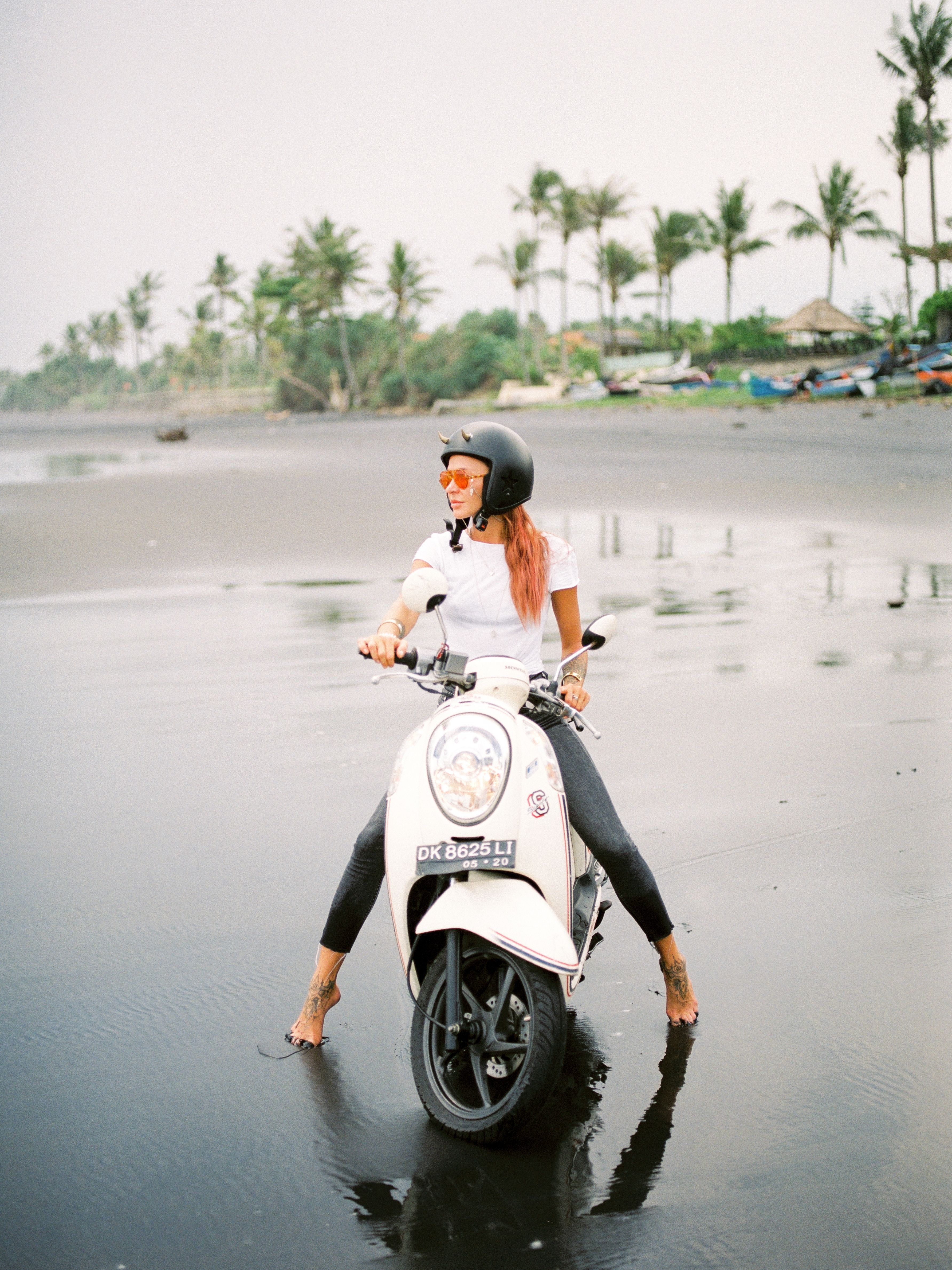A woman riding a scooter along a sandy Bali beach lined with palm trees